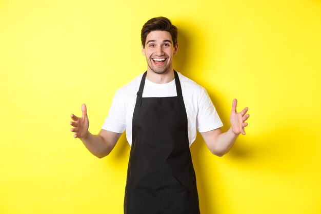 Feliz barista segurando algo grande, dando forma a um grande objeto, de pé sobre um fundo amarelo.