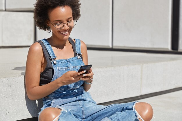 Feliz blogueira feminina na moda senta-se de pernas cruzadas do lado de fora, envia mensagem de texto para postar em site pessoal, está de bom humor, envia feedback, usa roupas elegantes para a juventude, aproveita o tempo de recreação