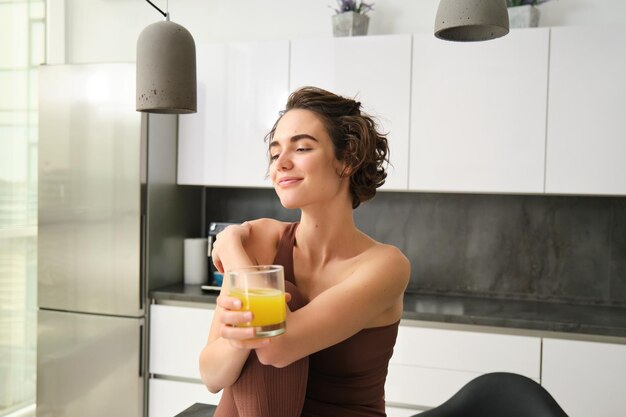 Feliz bruneta desportista garota em roupas de atividade bebendo um copo de suco de laranja sentada em sua cozinha