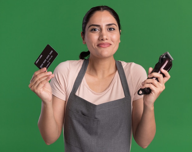 Feliz cabeleireira jovem bonita com avental segurando o aparador e o cartão de crédito, olhando para a frente com um sorriso no rosto em pé sobre a parede verde