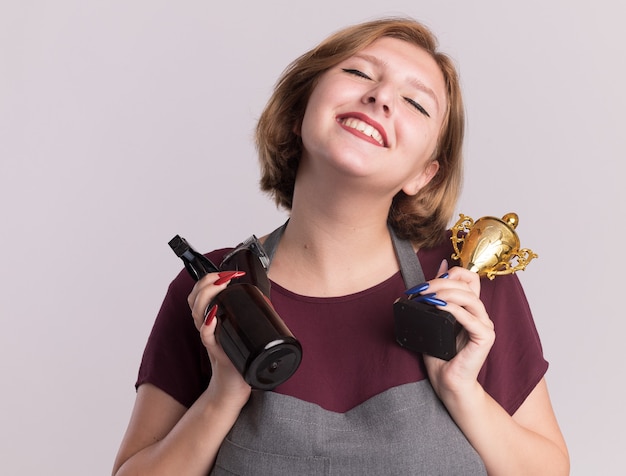 Feliz cabeleireira jovem bonita com avental segurando um troféu de ouro e um frasco de spray com aparador sorrindo com os olhos fechados em pé sobre uma parede branca