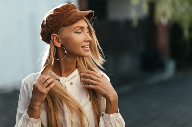 Feliz, calma, jovem loira bronzeada de boné marrom e camisa branca, sorrindo sinceramente e fazendo poses lá fora