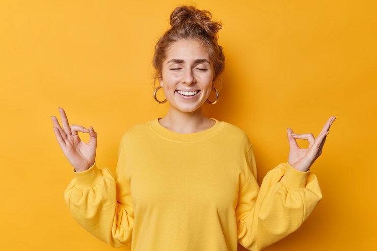 Foto grátis feliz calma paciente jovem faz gesto zen nirvana medita com os olhos fechados pratica ioga tem cabelo penteado usa brincos e sorrisos de jumper casual feliz isolado sobre fundo amarelo.