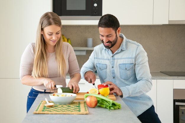 Feliz casal jovem atraente cozinhando o jantar juntos, cortando legumes frescos na tábua na cozinha, sorrindo e conversando. Conceito de cozinha familiar