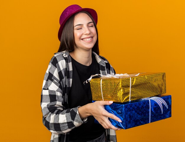 Feliz com os olhos fechados, uma jovem linda com um chapéu de festa segurando caixas de presente isoladas na parede laranja
