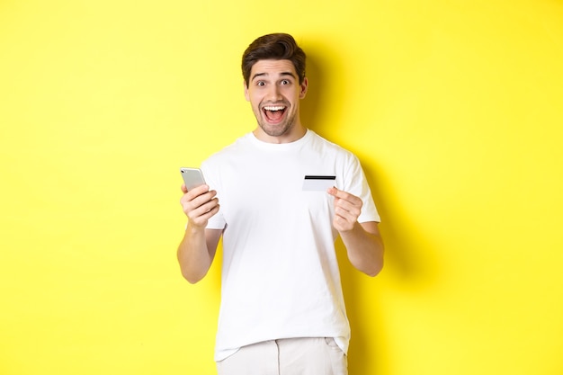 Feliz comprador masculino segurando smartphone e cartão de crédito, conceito de compras online na internet, em pé sobre fundo amarelo.