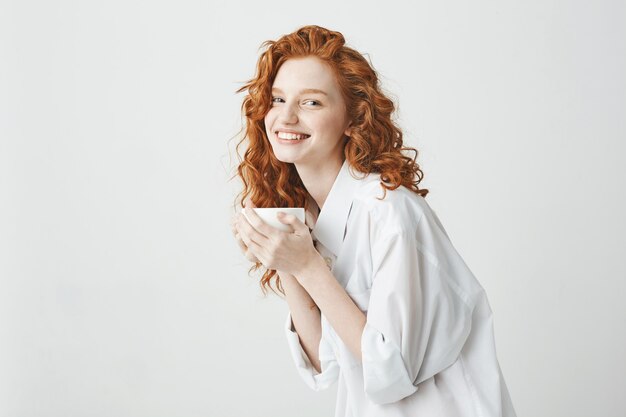 Feliz concurso garota ruiva na camisa sorrindo segurando copo sentado na mesa