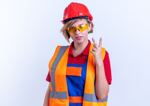 feliz construtora jovem de uniforme com óculos, mostrando gesto de paz isolado na parede branca