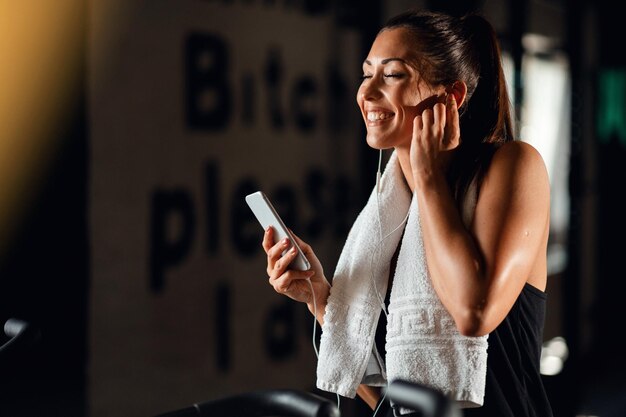 Feliz desportista usando telefone inteligente e curtindo boa música em fones de ouvido com os olhos fechados em uma academia