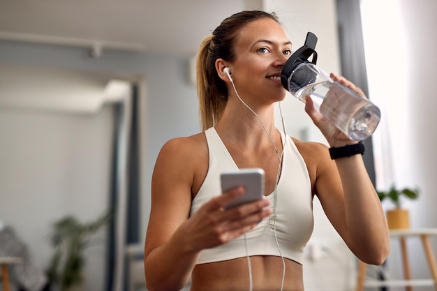 Feliz desportista usando telefone inteligente enquanto faz pausa para água na sala de estar