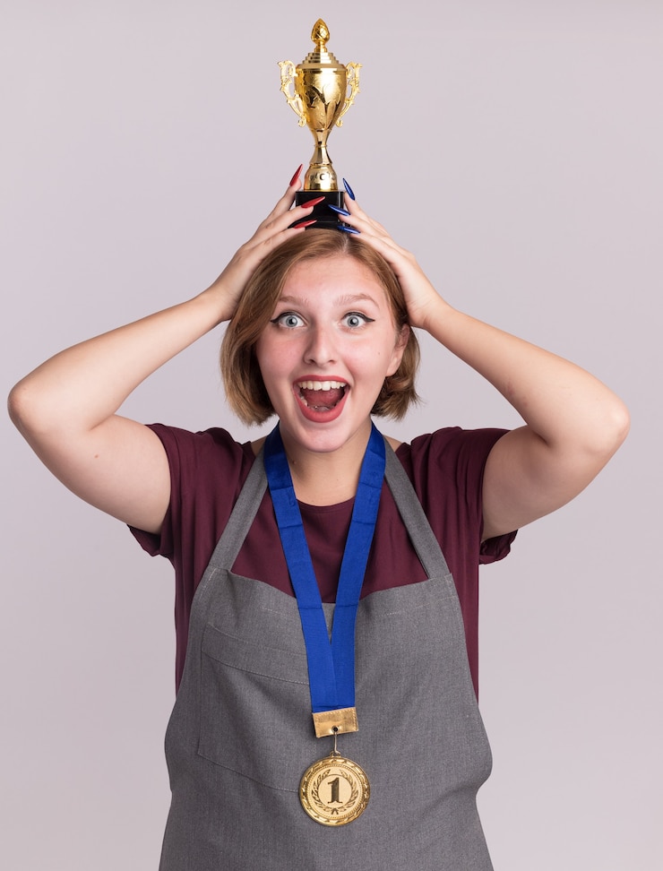 Foto grátis feliz e alegre cabeleireira jovem bonita com avental com medalha de ouro em volta do pescoço segurando um troféu de ouro na cabeça, olhando para a frente com um grande sorriso no rosto em pé sobre uma parede branca
