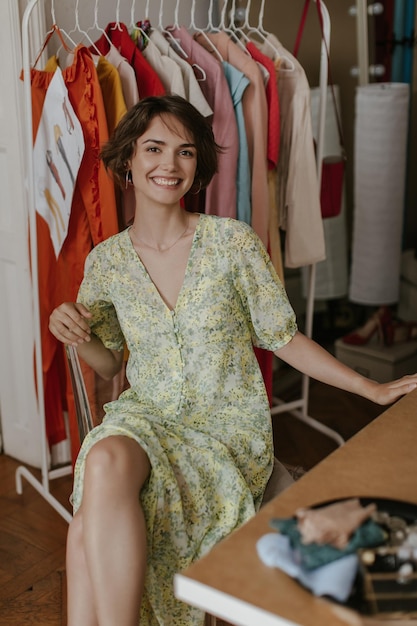 Foto grátis feliz e animada morena de cabelos curtos em vestido floral estiloso sorri sinceramente se senta na cadeira e posa no camarim