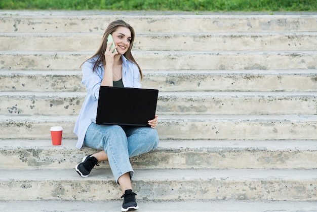 Feliz e atraente garota caucasiana está sentado com um laptop nas escadas e falando ao telefone. trabalho à distância. conceito freelance