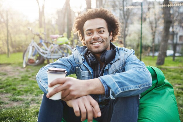 Feliz e positivo jovem estudante do sexo masculino com penteado afro em roupas da moda, sentado no parque, sorrindo amplamente e bebendo café.