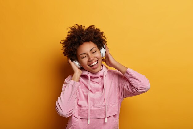 Feliz e relaxada melomana ouve música através de fones de ouvido, fecha os olhos e se sente otimista, vestida com um moletom casual, gosta de um som agradável, posa contra a parede amarela. Pessoas, lazer, felicidade