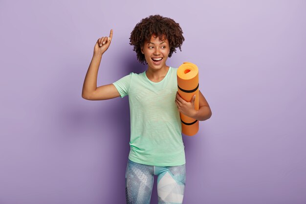 Feliz e sorridente desportista afro-americana de cabelos cacheados encaracolados carrega um tapete de ioga amassado, levanta o braço e indica para cima, desfruta de um bom treino, vestida com t-shirt e leggings. Conceito de esporte