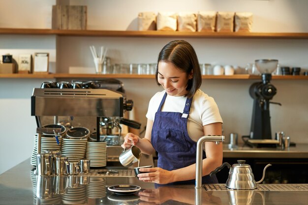 Feliz e sorridente proprietária de café barista em avental fazendo cappuccino latte art com standin de leite cozido no vapor