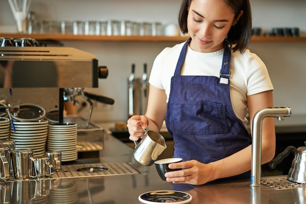 Feliz e sorridente proprietária de café barista em avental fazendo cappuccino latte art com standin de leite cozido no vapor