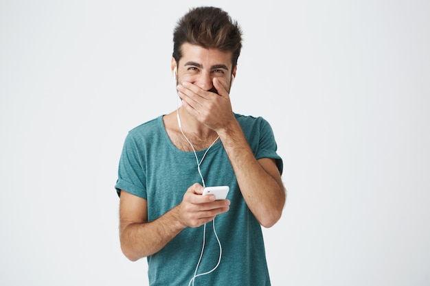 Foto grátis feliz elegante hispânico jovem vestindo camiseta azul elegante, sorrindo e cobrindo a boca com a mão, tentando não rir em voz alta no transporte.