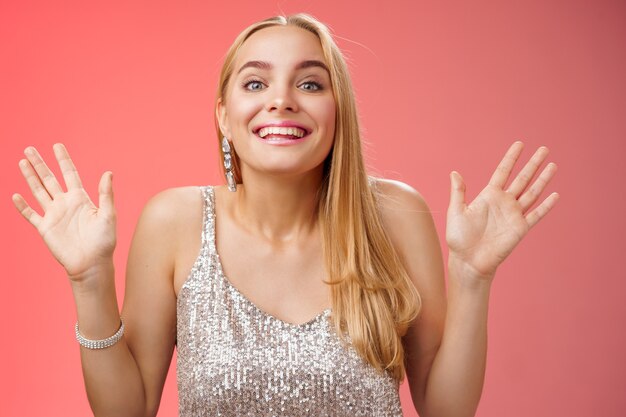 Feliz, elegante, sonhadora, glamour, jovem, mulher loira, levantando as mãos, sorrindo alegremente para a câmera, feliz por ver amigos chegando na festa dando as boas-vindas aos convidados sorrindo alegremente, usando um vestido prateado elegante