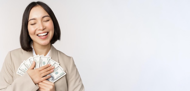 Feliz empresária asiática segurando dinheiro abraçando dólares e sorrindo em pé sobre fundo branco