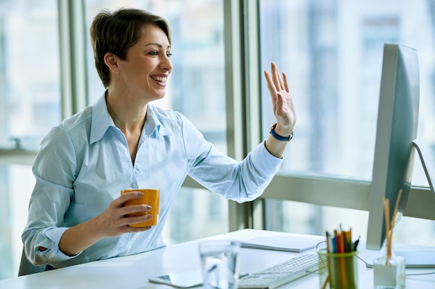 Feliz empresária fazendo videochamada no computador enquanto toma café no escritório