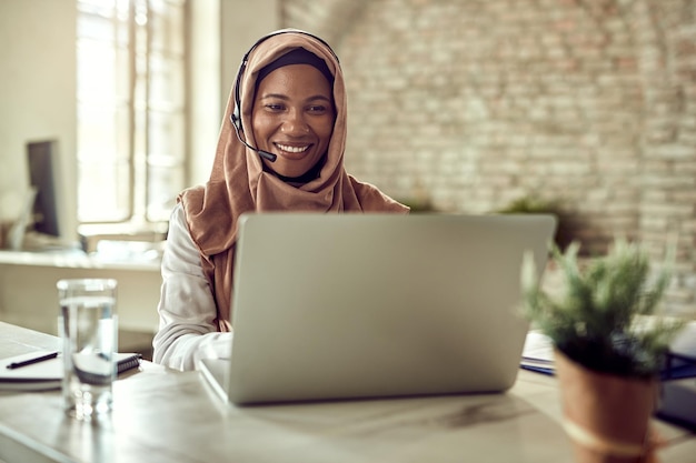 Feliz empresária islâmica trabalhando em um computador e se comunicando por fone de ouvido no escritório