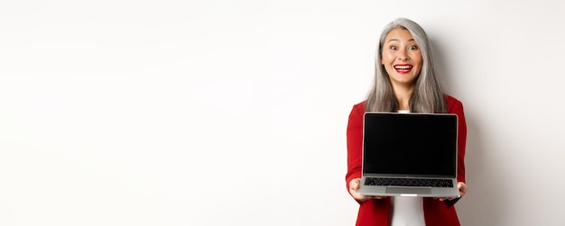 Feliz empresária sênior de negócios demonstra tela de laptop em branco sorrindo espantada com a câmera s