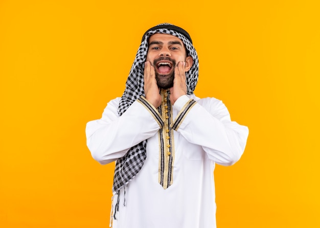 Feliz empresário árabe com roupas tradicionais e sorrindo alegremente em pé sobre a parede laranja