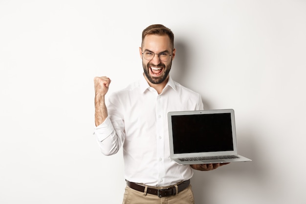 Feliz empresário mostrando a tela do laptop, levantando o punho e regozijando-se com a conquista online, em pé