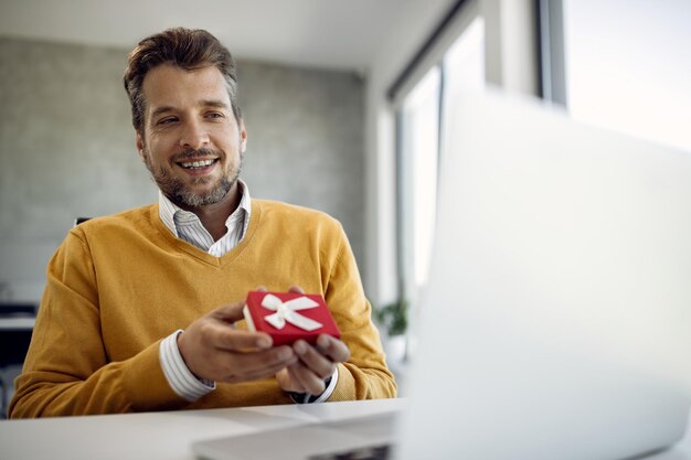 Feliz empresário segurando uma pequena caixa de presente enquanto faz videochamada no laptop