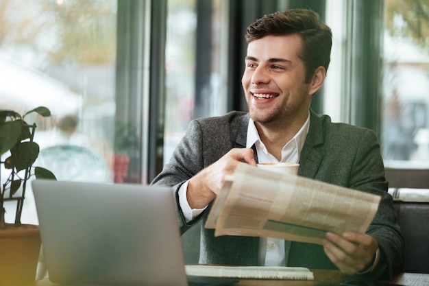 Foto grátis feliz empresário sentado junto à mesa de café com computador portátil e jornal enquanto bebia café e desviar o olhar