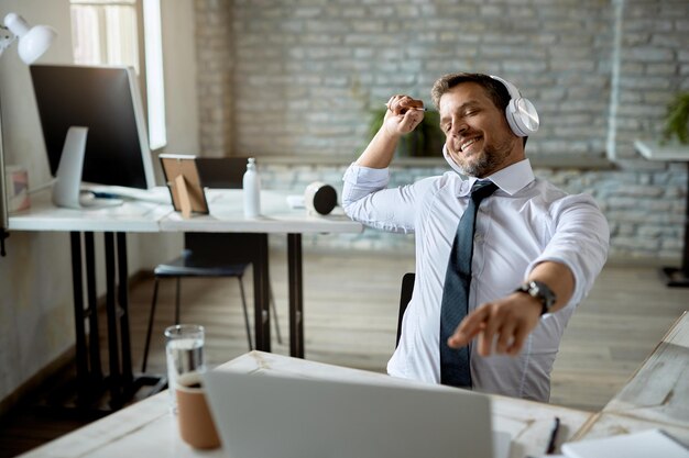 Feliz empresário usando computador e se divertindo enquanto ouve música em fones de ouvido no escritório