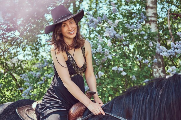 Feliz encantadora linda morena vestindo roupas pretas e chapéu montando um cavalo marrom no jardim de flores.