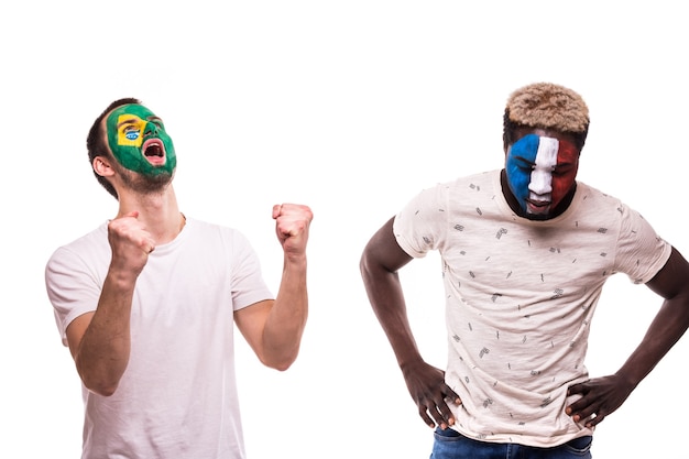 Foto grátis feliz fã de futebol do brasil comemora vitória sobre chateado fã de futebol das seleções francesas com o rosto pintado isolado no fundo branco