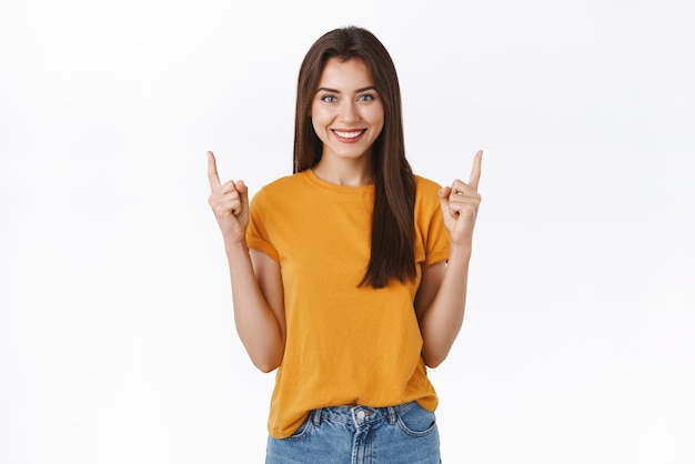 Feliz feliz e simpática jovem morena de camiseta amarela, levantando os dedos e apontando para cima sorrindo confiante, com certeza de que você gostará de nova promoção recomendo clique no link ou participe do evento