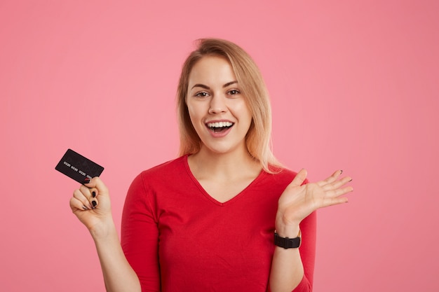 Feliz fêmea loira animado detém cartão de plástico, não espera receber salário, vai fazer pagamentos, olha alegremente para a câmera