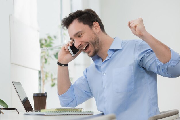 Feliz freelancer rindo enquanto fala pelo celular Homem bonito de camisa azul trabalhando no laptop dentro de casa