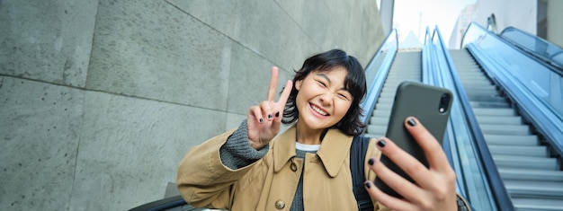 Foto grátis feliz garota asiática triunfante parece surpresa e alegre segura smartphone descendo escada rolante com