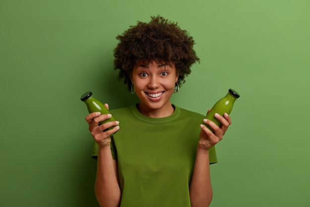 Foto grátis feliz garota de pele escura segura garrafas de vidro com smoothie vegetal de desintoxicação verde cru, leva um estilo de vida saudável, mantém a dieta vegetariana, se sente revigorada e feliz. foto monocromática. pessoas e bem-estar