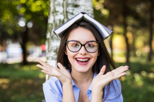 Feliz garota séria de cabelos escuros de jaqueta jeans e óculos lê um livro contra o parque verde de verão.