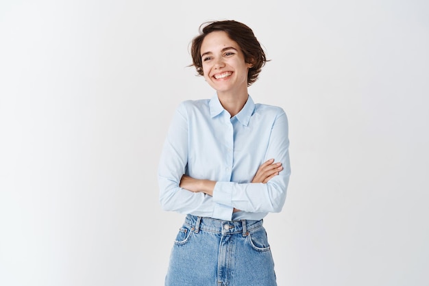 Feliz gerente feminina sincera de camisa azul e jeans cruzam os braços no peito olhando de lado e sorrindo alegremente em pé no fundo branco