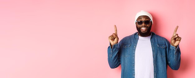 Feliz homem africano-americano barbudo mostrando anúncio apontando dedos para cima e sorrindo em pé em b