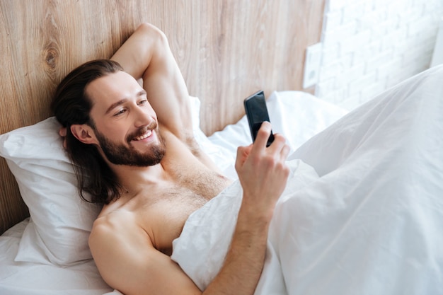 Feliz homem barbudo, deitado na cama e usando telefone celular