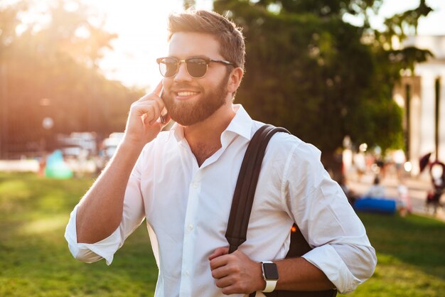 Feliz homem barbudo em óculos de sol em pé ao ar livre, segurando a mochila e falando pelo smartphone