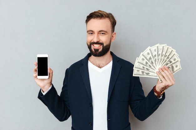 Feliz homem barbudo em roupas de negócios, mostrando dinheiro e smartphone