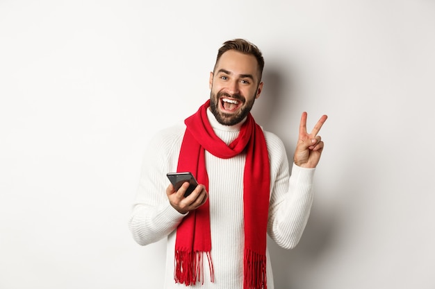 Feliz homem barbudo usando smartphone, posando para uma foto com o símbolo da paz, em pé na camisola de inverno e lenço vermelho, fundo branco.