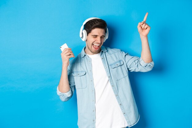 Feliz homem bonito dançando, ouvindo música no smartphone em fones de ouvido, em pé sobre um fundo azul