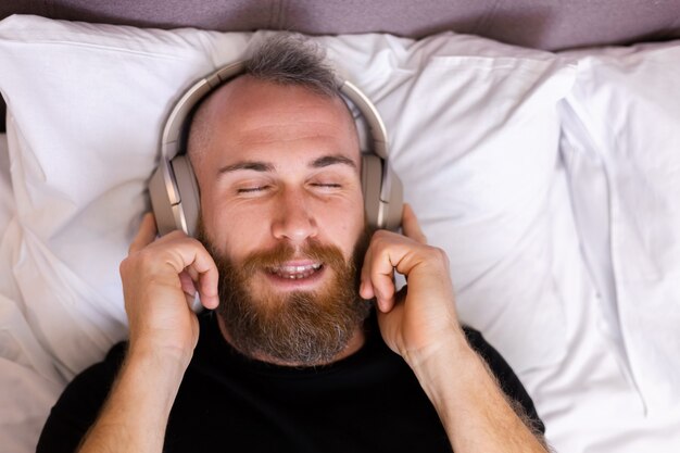 Feliz homem caucasiano na cama usando fone de ouvido ouve curtir sua música favorita, descansando sozinho, dançando.