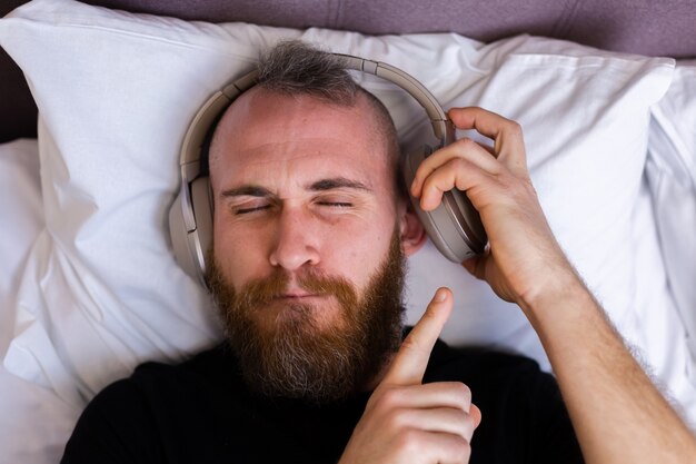 Feliz homem caucasiano na cama usando fone de ouvido ouve curtir sua música favorita, descansando sozinho, dançando.